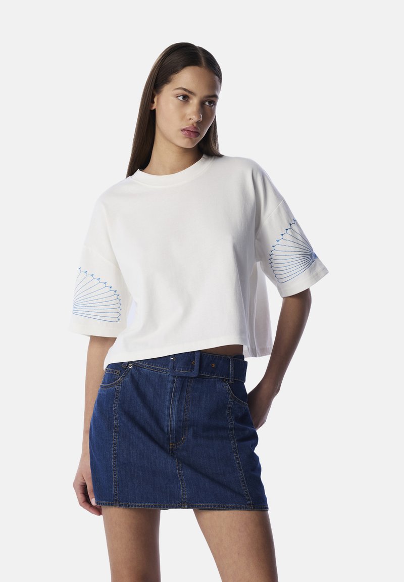 Witte cropped t-shirt met blauwe borduursels op de mouwen, gecombineerd met een donkerblauwe denim mini-jurk met een frontale knoopsluiting en riem.