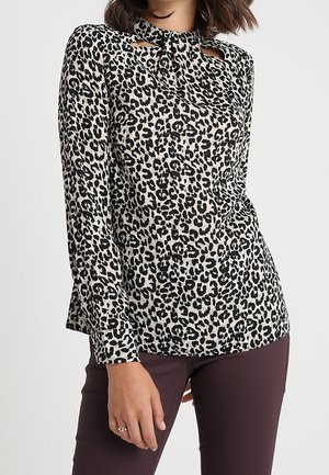 Kobieta ubrana w długą bluzkę z nadrukiem w czarno-białe cętki lamparta, z wysokim kołnierzem i wycięciami w kształcie łezki, zestawioną z ciemnymi spodniami.