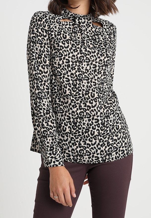 Femme portant un chemisier à manches longues, imprimé léopard noir et blanc, avec un col montant et des découpes en forme de goutte, assorti à un pantalon foncé.