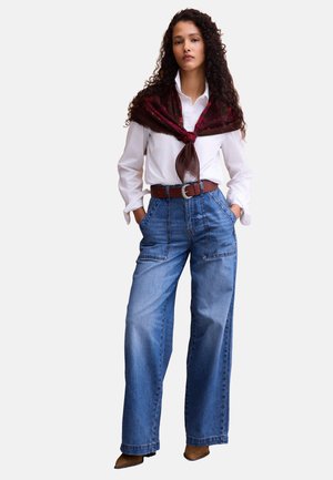 Frau mit lockigem Haar, die ein weißes Hemd, einen braunen Schal über die Schultern gelegt, blaue Jeans mit hoher Taille und weitem Bein, einen braunen Gürtel und braune spitze Schuhe trägt.