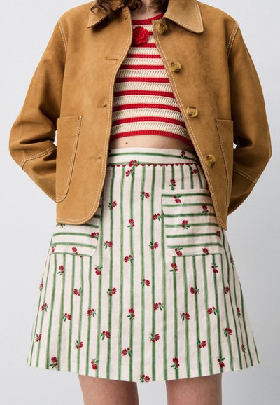Giacca in suede marrone con cuciture visibili, top corto a righe rosse e crema, e gonna bianca con strisce verdi e ricamo di fiori rossi.