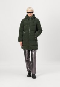 Chaqueta de plumas verde con tejido texturizado, cremallera frontal, capucha y dos bolsillos laterales. Combinada con pantalones plateados brillantes y zapatos negros.