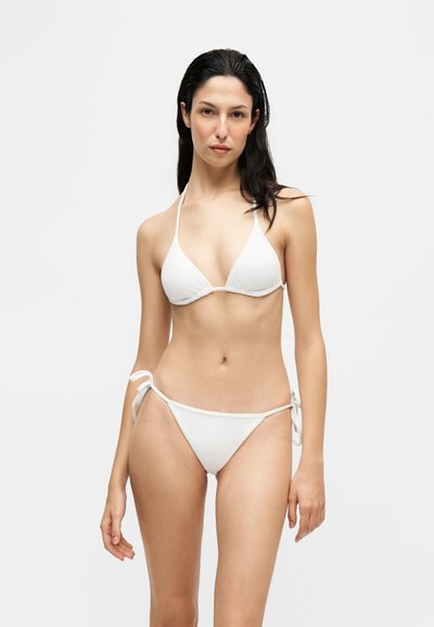 Femme aux longs cheveux foncés portant un bikini string blanc, se tenant devant un fond clair uni, regardant droit devant avec une expression neutre.