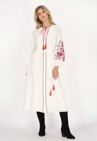 Weißes langärmliges Kleid mit roten gestickten Blumenmustern, Kordelzug am Hals, Taillengürtel und Stoffquasten. Getragen mit schwarzen Stiefeln mit Absatz.