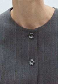 Grijze pinstriped blazer met een ronde hals, voorzien van twee zwarte knopen en een gestructureerde stof. De knopen zijn centraal geplaatst onder de hals.