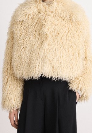 Beżowa krótka kurtka z kręconego sztucznego futra noszona na czarnych, wysokich spodniach, z jedną ręką w kieszeni i neutralnym tłem.