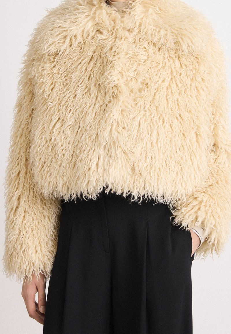 Veste courte en fausse fourrure bouclée beige portée sur un pantalon noir taille haute, une main dans la poche, sur un fond neutre.