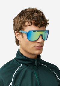Siniset ylisuurikokoiset aurinkolasit, joissa on liukuvärjätty peiliobjektiivi, urheilullinen muotoilu ja erottuva kulmikas muoto, käytetty vihreän vetoketjullisen takin kanssa.