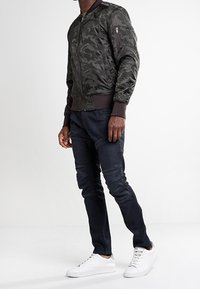 Man som bär en mörk kamouflage bomberjacka, svarta slim-fit byxor och vita sneakers, stående mot en helt vit bakgrund.