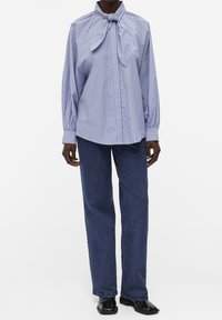 Camicia a righe blu e bianche con colletto a fiocco, maniche lunghe e davanti con bottoni. Abbinata a jeans in denim scuro e scarpe nere.