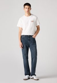 Camiseta branca de mangas curtas com um gráfico colorido no peito, combinada com jeans de lavagem média em azul e ténis brancos com riscas pretas.