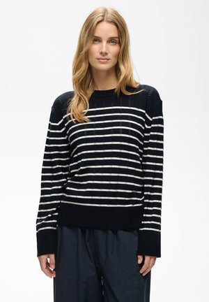 Femme aux longs cheveux blonds portant un pull rayé bleu marine et blanc et un pantalon ample bleu marine, debout devant un fond uni clair.