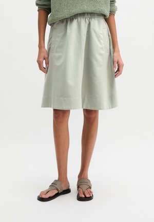 Kobieta ubrana w jasnooliwkową spódnicę do kolan z bocznymi kieszeniami, zielony sweter dzianinowy oraz taupe sandały z czarną podeszwą.