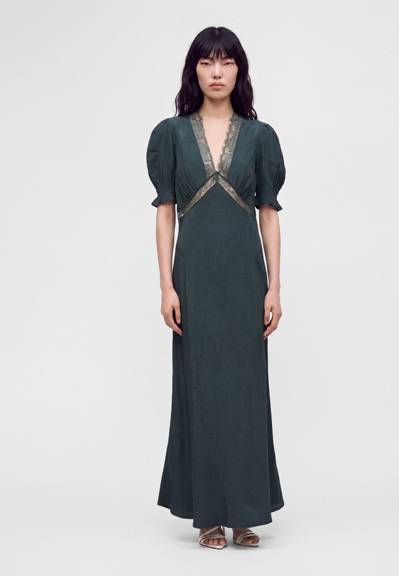 Femme aux longs cheveux noirs debout, portant une robe longue vert foncé avec une bordure en dentelle et des manches courtes bouffantes, ainsi que des sandales blanches.