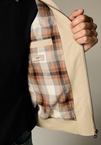 Main tenant une veste beige ouverte montrant la doublure intérieure à carreaux aux tons marron et crème ainsi qu'une poche intérieure étiquetée "NEXT".