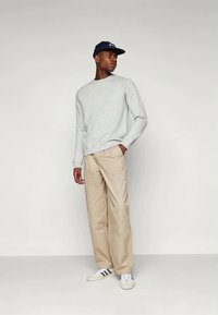 Hombre vestido con un suéter gris claro, pantalones beige, gorra negra y zapatillas blancas con rayas negras, de pie contra un fondo claro y liso.