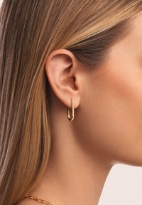 Boucle d'oreille en or avec un design lisse et courbé, ornée d'une rangée de petits cristaux le long d'un bord. Portée sur une oreille avec des cheveux lisses et châtain clair.