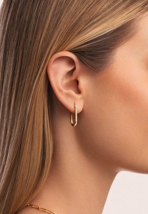 Pendiente de aro de oro con un diseño curvado y suave que presenta una fila de pequeños cristales a lo largo de un borde. Lleva puesto en una oreja con cabello lacio y castaño claro.