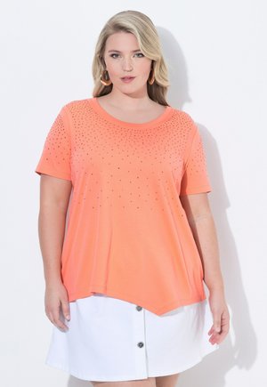 Blonde vrouw draagt oranje korte mouw top met studs en witte rok met knopen, staand tegen een witte achtergrond.