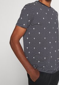 Granatowa koszulka z krótkimi rękawami w białe poziome paski i małe wzory palm. Design z okrągłym dekoltem i luźny krój. Miękka faktura materiału.