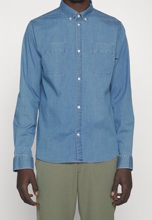 Homme portant une chemise en denim bleue à boutons avec poches poitrine et un pantalon vert olive, debout devant un fond blanc uni.