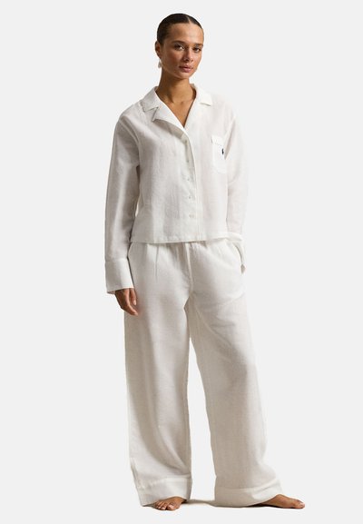 Jonge vrouw staat op blote voeten, draagt een loszittend wit overhemd met kraag en bijpassende broek met brede pijpen tegen een effen achtergrond.