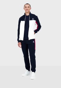 Träningsdräkten har en marinblå kropp med vita och vinröda detaljer, zip-up jacka, långa ärmar och figurnära byxor. Bärs med vita sneakers.