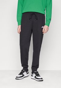 Calças de jogging pretas em algodão com cintura elástica e punhos canelados, apresentando um logótipo da Nike branco na coxa esquerda, combinadas com ténis pretos e brancos.