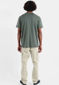 T-shirt en coton vert à encolure ronde, associé à un pantalon cargo beige avec poches latérales et poignets élastiques, porté avec des chaussures noires.