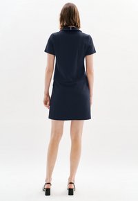 Abito navy a maniche corte con corpino aderente e gonna svasata, caratterizzato da un colletto sottile e una texture liscia. Vengono indossati sandali neri con tacco.