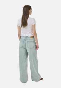 Woman with long brown hair wearing a white short-sleeve top and light blue wide-leg jeans, standing with back facing the camera.