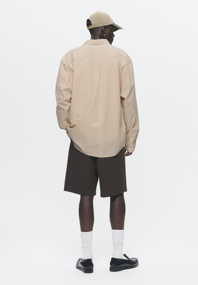 Chemise rayée à manches longues de couleur orange clair et blanche, associée à un short marron foncé. Les accessoires comprennent une casquette beige et des chaussettes blanches.