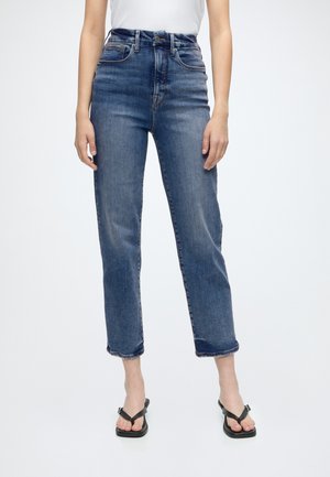 Femme portant un jean denim bleu taille haute avec poches avant et sandales noires à bout ouvert, debout devant un fond uni.