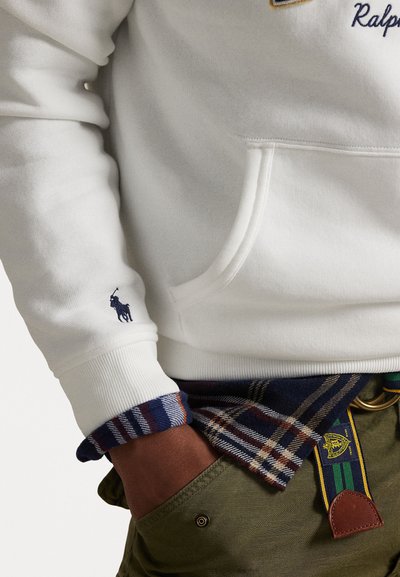 Felpa bianca con tasca a marsupio e polsini a coste, caratterizzata da un logo ricamato blu navy sulla manica. Sotto, una camicia a quadri con colori blu e marrone.