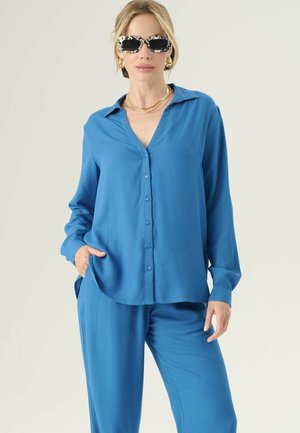 Camicia blu con colletto a V, maniche lunghe e vestibilità rilassata. Realizzata in tessuto morbido, presenta una leggera lucentezza e tasche laterali.