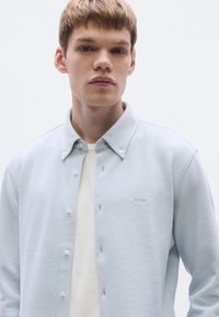 Svetlomodrá košeľa s golierom zo štruktúrovanej látky, s gombíkmi na prednej strane a malým logom na ľavej strane. Nosená cez bielu košeľu.