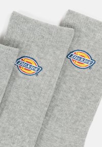 Pelēki adīti zeķes ar ribota tekstūra un logotipa uzšuvums, kurā ir vārds "Dickies" zilā krāsā uz apļa oranžā fona.