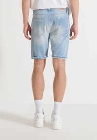 Pantalones cortos de mezclilla azul claro con dobladillo deshilachado, cinco bolsillos y un ligero desgaste. Lleva puestos unos zapatos deportivos blancos y calcetines blancos hasta el tobillo.