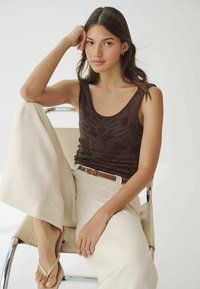 Jonge vrouw met lang haar zit op een stoel, gekleed in een bruine mouwloze top, beige wijde broek, bruine riem en beige sandalen.