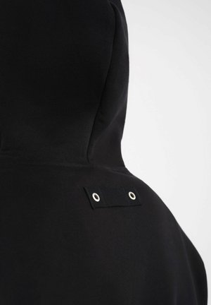 Zwarte hoodie achterkant met stoffen lus en twee zilveren ringen vlak bij het bovenste schoudergebied tegen een effen lichte achtergrond.