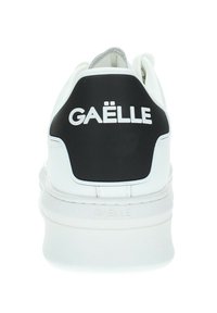 Sneaker bianche con un rialzo nera sul tallone etichettato "GÄËLLE." Materiale sintetico liscio, forma arrotondata, suola testurizzata e design minimalista.
