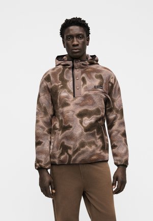 Homme portant un pull polaire camouflage marron avec capuche et boutons-pression, assorti d'un pantalon marron, debout devant un fond uni.