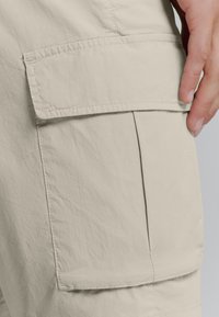 Beige cargobroek van een gladde, lichte stof. Heeft een grote opgestikte zak aan de zijkant en subtiele stiksel details.