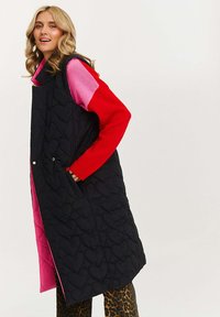 Wypełniona kamizelka w kolorze czarnym z wzorem serc, z różowym podszyciem, dwoma bocznymi kieszeniami i bezrękawnikowym krojem, noszona na czerwonym swetrze i wzorzystych spodniach.