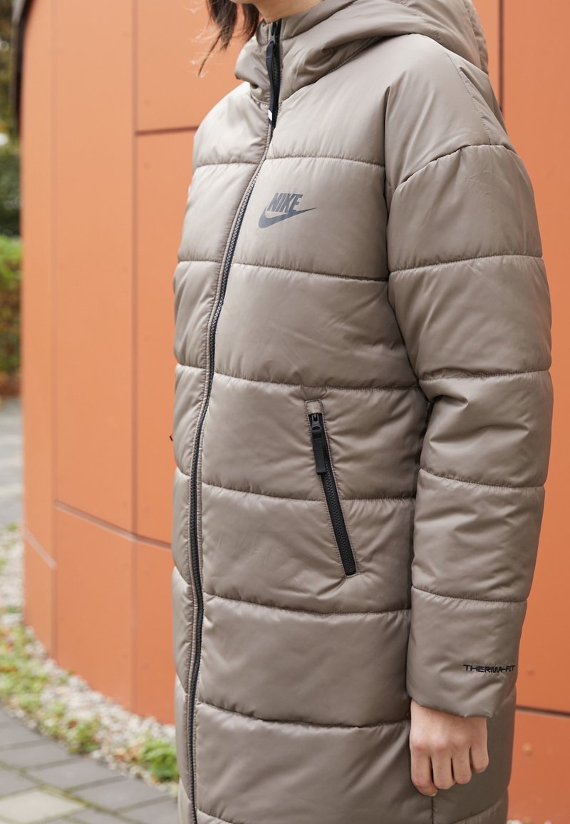 Padded long coat in khaki, featuring a zip closure, black accents, and a hood. Includes prominent Nike logo and Therma-Fit branding.