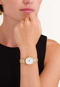 Zlatni ručni sat s bijelim brojčanikom, tankim oznakama sati i narukvicom od povezanih karika. Sat je nosen na ruci svijetle puti.