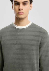 Tkaninowy sweter w oliwkowym kolorze z okrągłym dekoltem i białą podszywką przy kołnierzu. Ma poziomy wzór w paski i prążkowaną tkaninę.