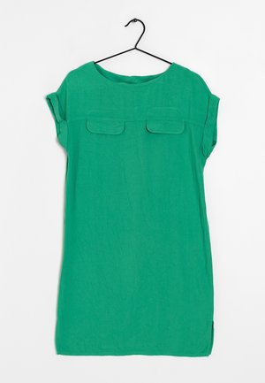 Zielona sukienka z krótkimi rękawami i klapkami na kieszeniach na piersiach, zawieszona na czarnym metalowym wieszaku na białym tle.