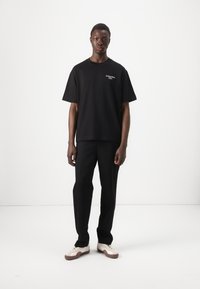 Black cotton t-shirt with a relaxed fit, short sleeves, and a small printed logo on the chest, paired with black trousers and white sneakers.