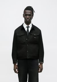 Schwarze Jeansjacke mit zwei Brusttaschen, Knopfleiste und Kragen. Kombiniert mit einem weißen gestreiften Hemd und schwarzer Krawatte. Dunkle Jeans.